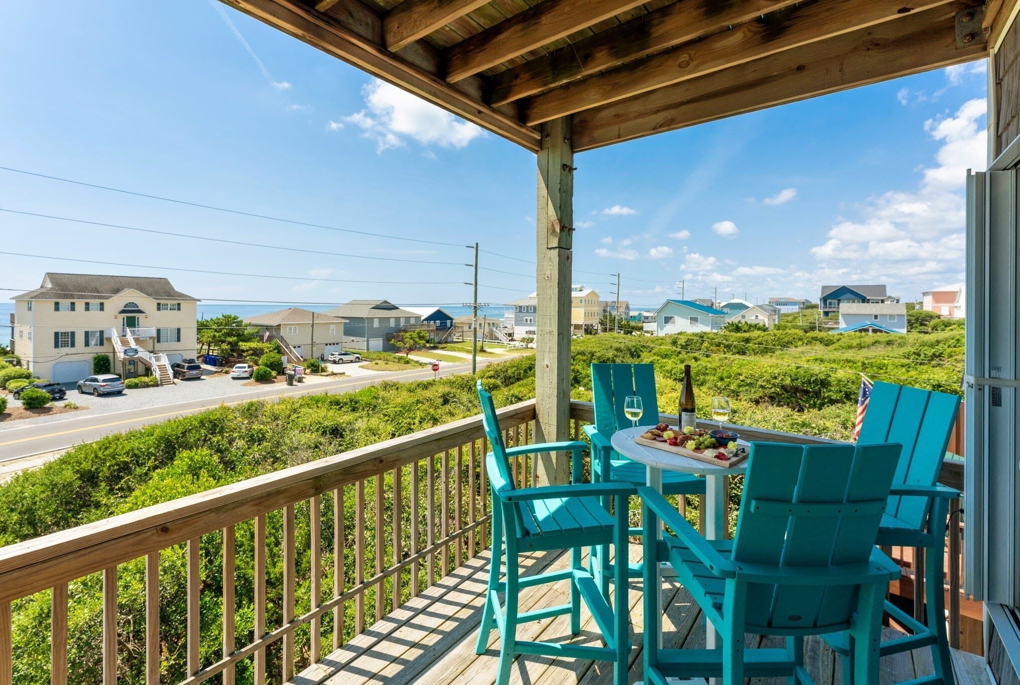 A wooden deck with turquoise chairs and a table set for dining, overlooking a residential area with houses and a road.