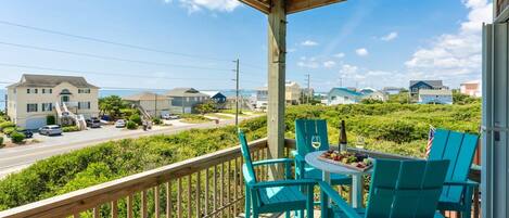A wooden deck with turquoise chairs and a table set for dining, overlooking a residential area with houses and a road.