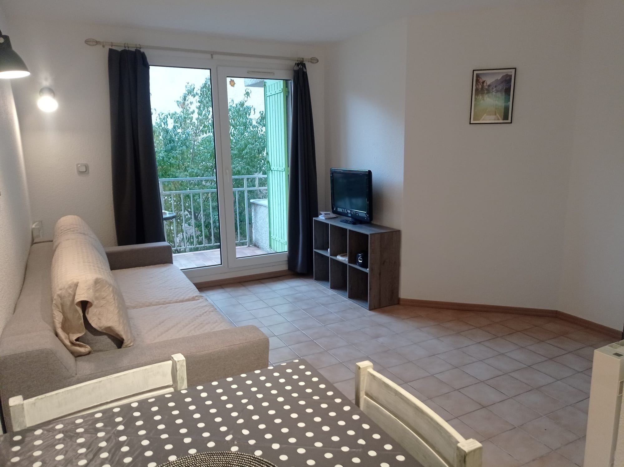 A room with a sofa, a TV on a wooden stand, a balcony with a view of greenery, and a painting on the wall.