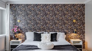 Une chambre à coucher avec un papier peint à motifs floraux, un lit avec une literie blanche et noire, deux tables de chevet avec des lampes et un vase rempli de fleurs.