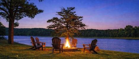 A lakeside setting with a fire pit and Adirondack chairs.