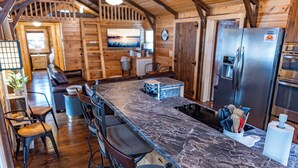 A wooden cabin interior with a kitchen area featuring a marble countertop, a refrigerator, and a dining area with a sofa and chairs. - Waterfront Retreat for 8 w/Fire Pit, Boat Dock, no steps to water! 3BR/2BA (Cadiz)