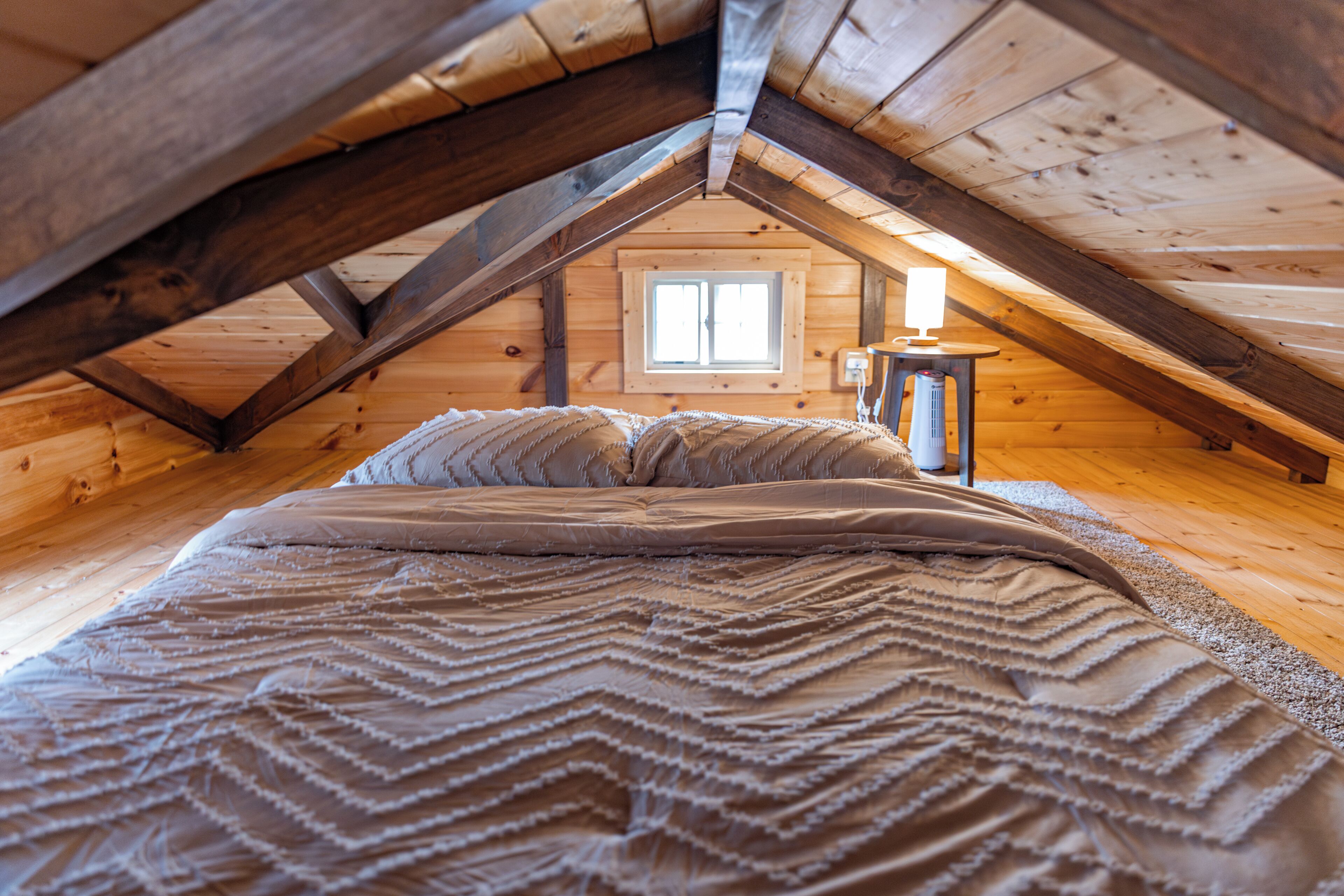 A cozy attic bedroom with a wooden ceiling, a bed with a patterned blanket, and a small table with a lamp.