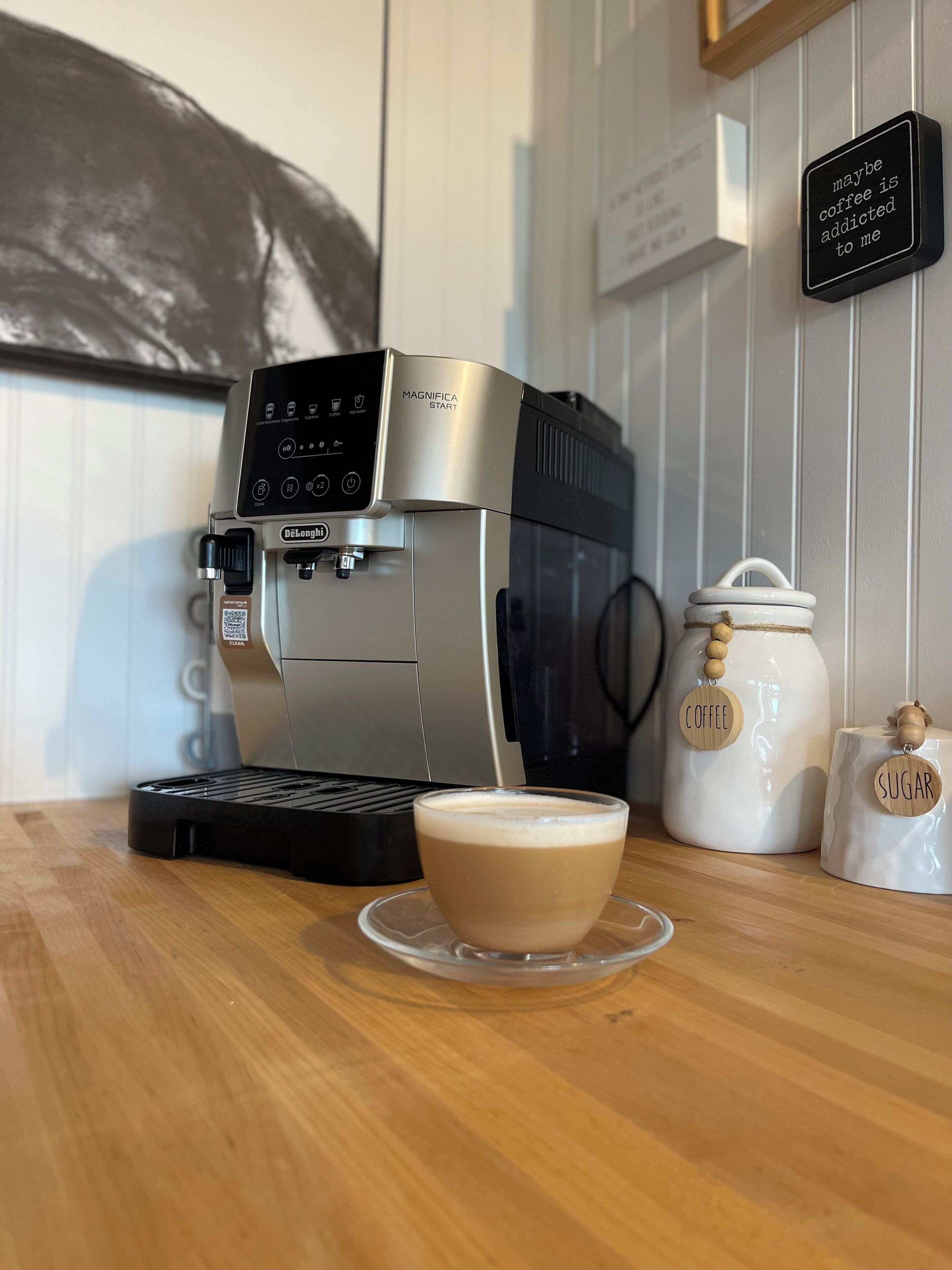 Una cafetera con una taza de café sobre una mesa de madera, junto a frascos etiquetados como 'Coffee' y 'Sugar'.