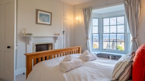 A bedroom with a wooden bed, white bedding, a fireplace, a window with curtains, and a view of buildings. - Spacious, cosy 5 bedroom home in the village of Seaview on the Isle of Wight views across the solent (Seaview)