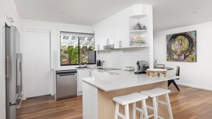 A modern kitchen with white cabinetry, a dishwasher, a coffee machine, and a small island with stools. - Solace by the Bay (Corlette)
