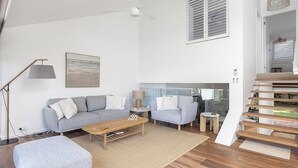 A modern living room with a grey sofa, a wooden coffee table, and a floor lamp. There is a staircase with wooden steps leading to an upper level. - Solace by the Bay (Corlette)