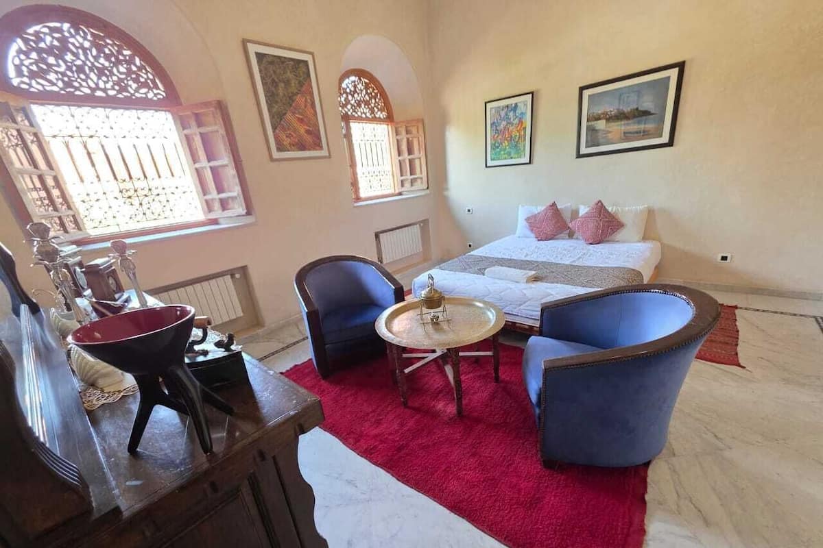 A hotel room with a bed, two blue armchairs, a small round table, a red rug, and a decorative wall.