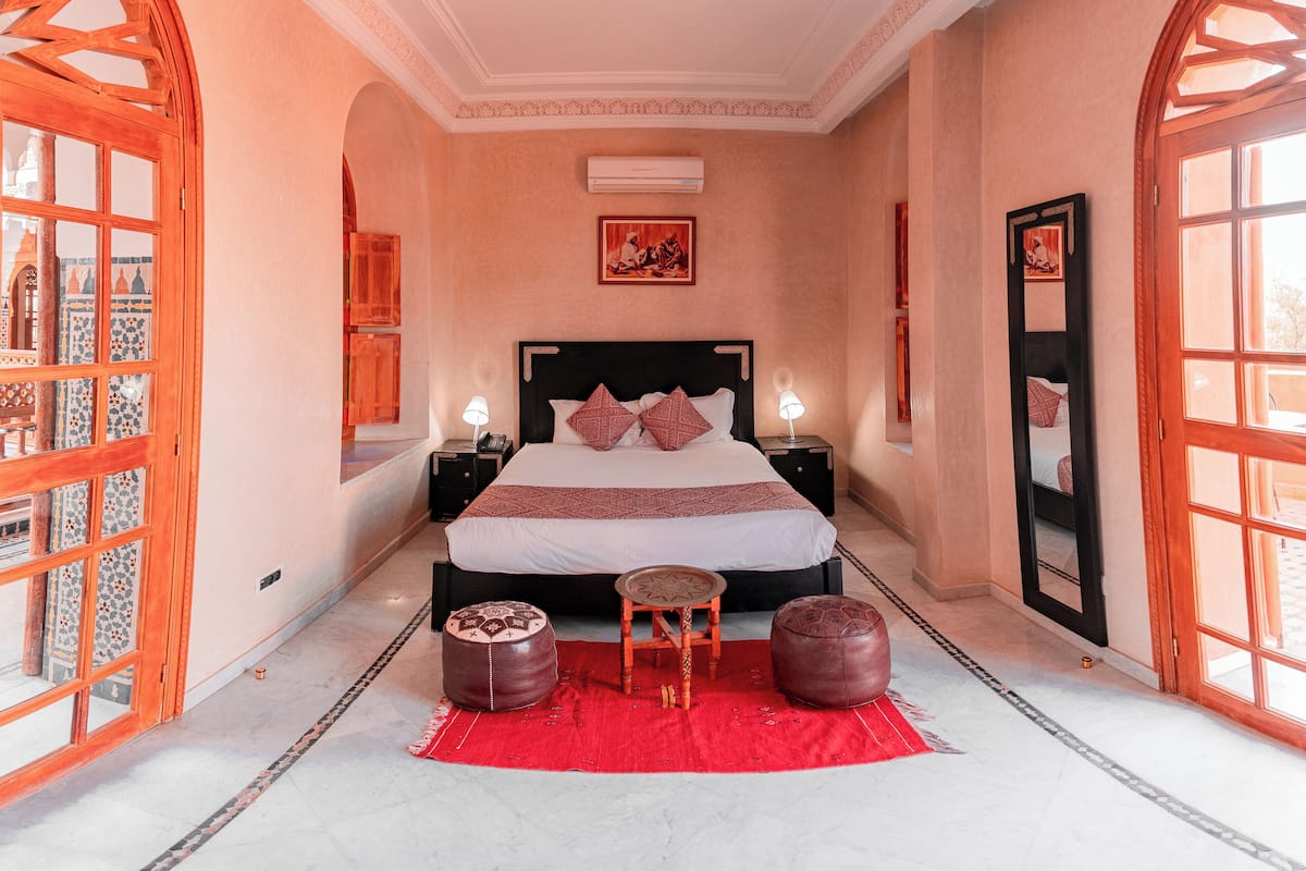 A spacious bedroom with a large bed, two nightstands, a red rug, and a large window with wooden frames.