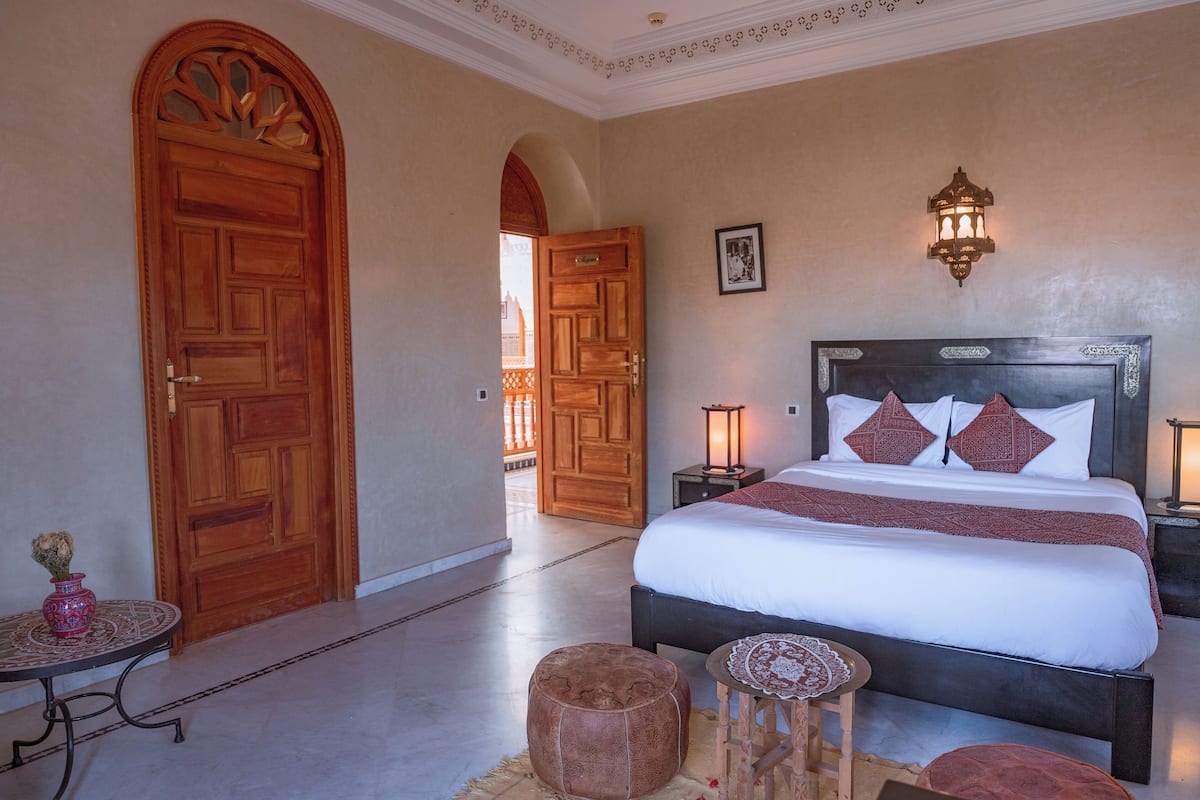A hotel room with a large bed, two bedside tables with lamps, a small round table, and a decorative stool.