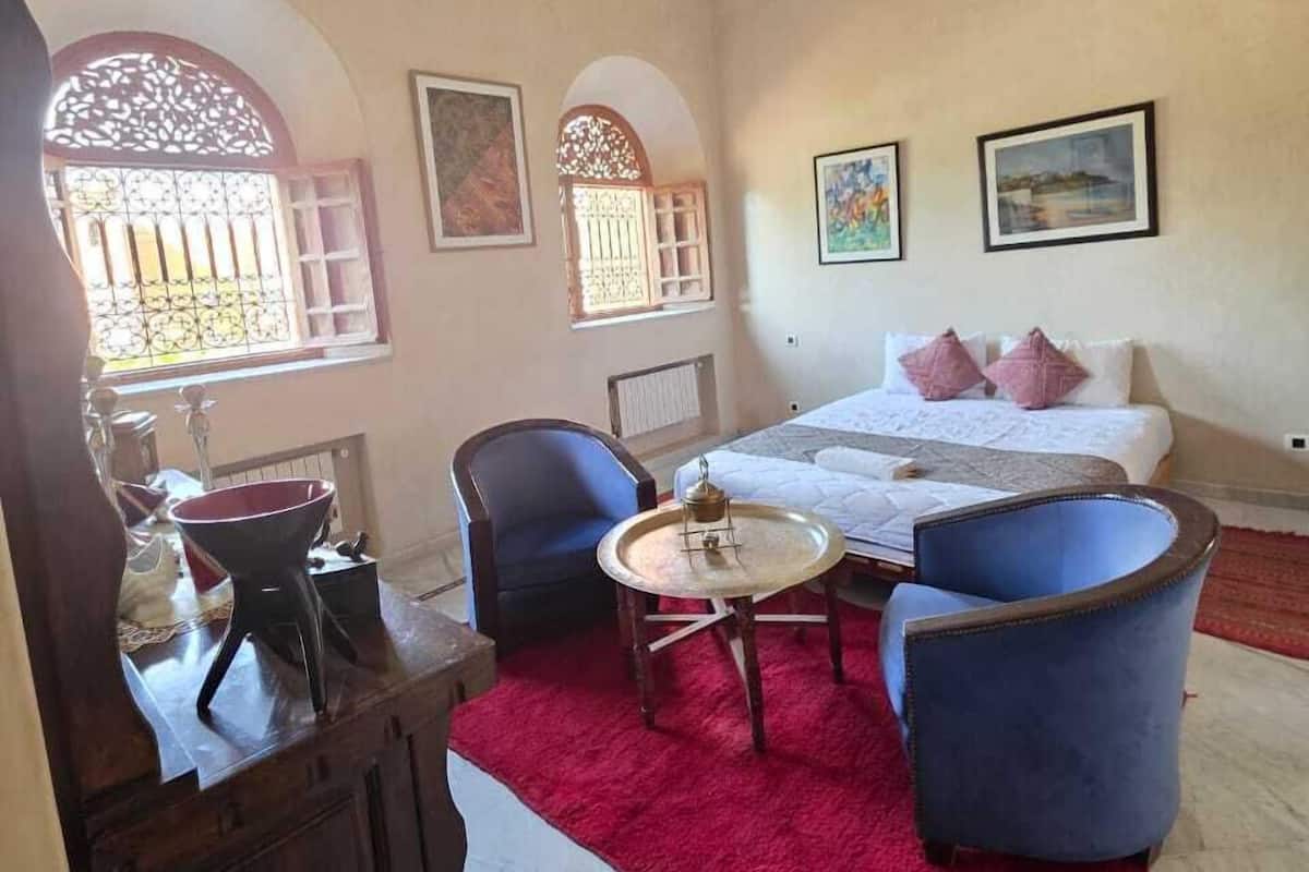 A bedroom with a bed, two chairs, a small table, a chandelier, and artwork on the walls.