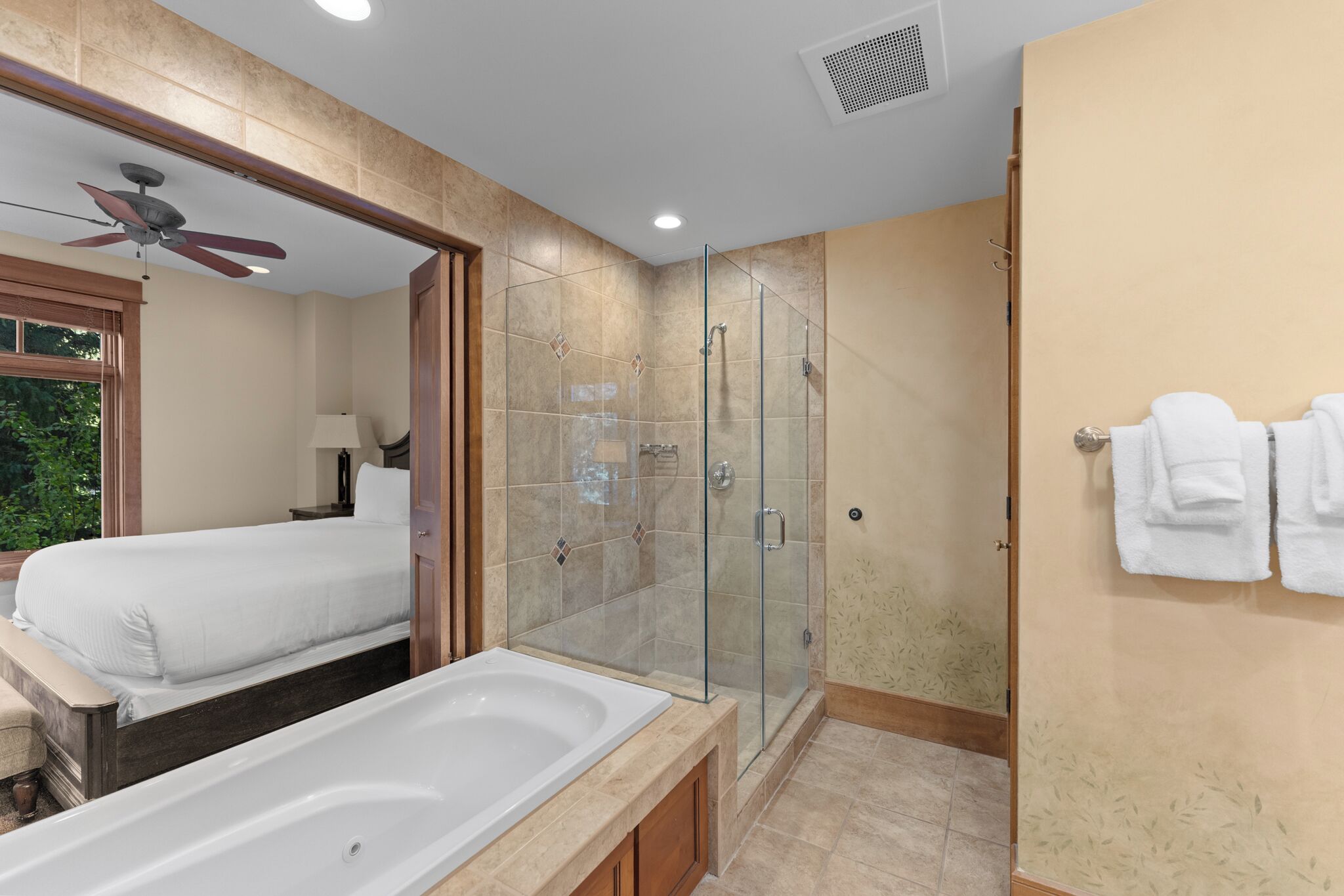 A modern bathroom with a glass-enclosed shower, a large bathtub, and a bed with a ceiling fan.