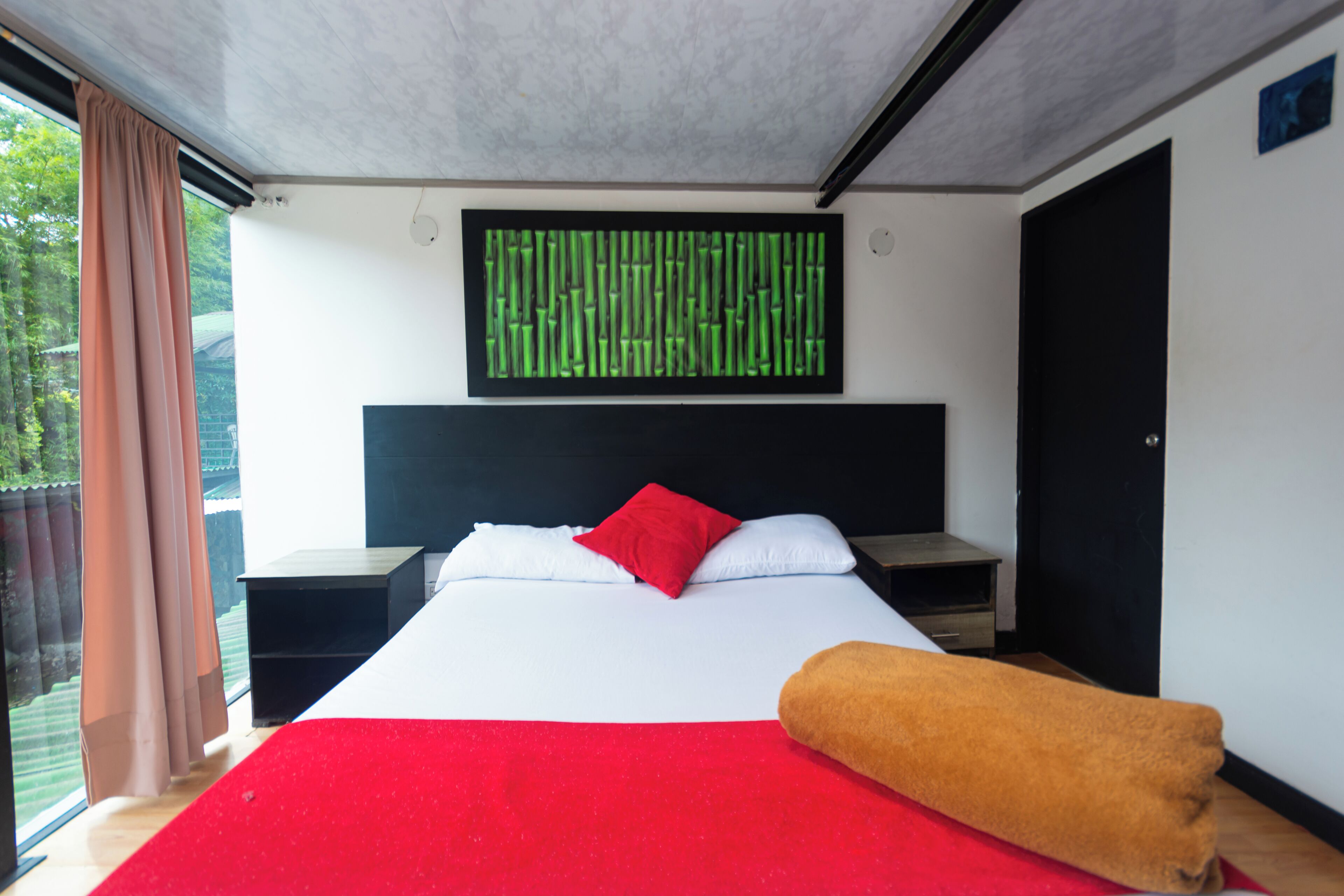A modern hotel room with a large bed, a red and white bedspread, a red pillow, a brown throw blanket, a black headboard, a nightstand, a small table, a wall-mounted TV, and a window with curtains.