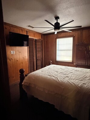 Een slaapkamer met een bed, houten wanden, een plafondventilator, een raam met jaloezieën en een tv aan de muur.