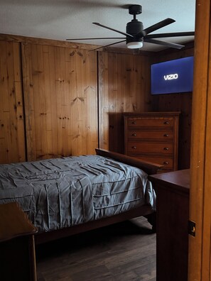 Une chambre à coucher comprenant un lit, une commode en bois, un ventilateur de plafond et un téléviseur fixé au mur.