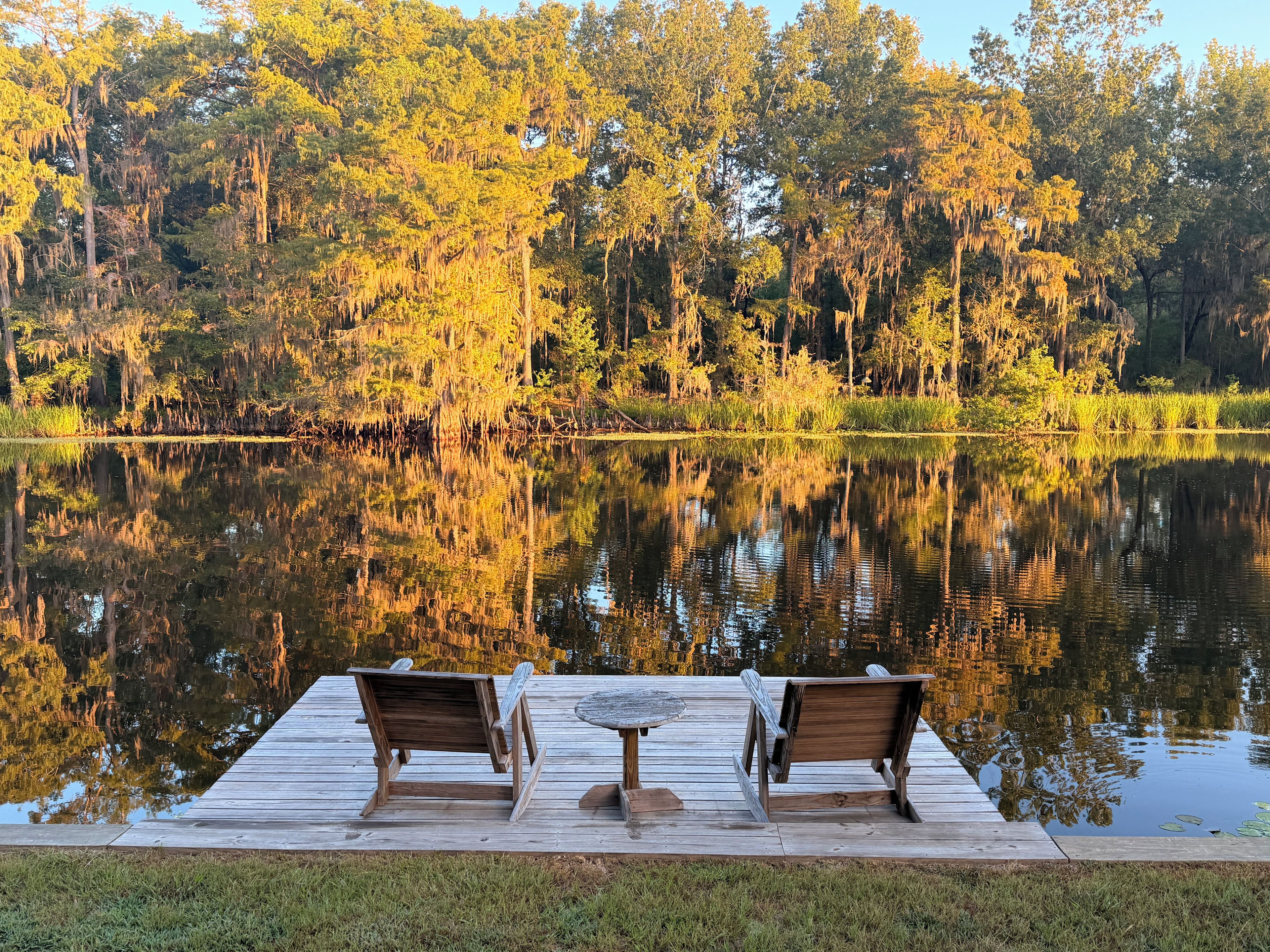 Una terraza de madera con dos sillas y una mesa con vista a un lago tranquilo, cuyas aguas reflejan árboles y cielo.