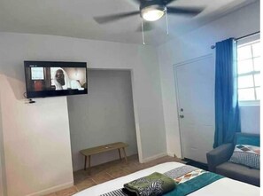 A hotel room with a bed, a bench, a television mounted on the wall, and a door with a blue curtain. - Spacious two bedrooms in Providenciales (The Bight Settlements)