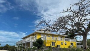 A yellow two-story house with a balcony, surrounded by a stone wall and vegetation. - Waterfront 2-BR Home in Providenciales (Turtle Cove)