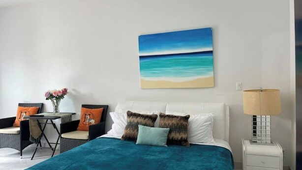 Ein Hotelzimmer mit Bett, Nachttisch, Lampe, Sessel, kleinem Tisch und einem Bild von einer Strandlandschaft.