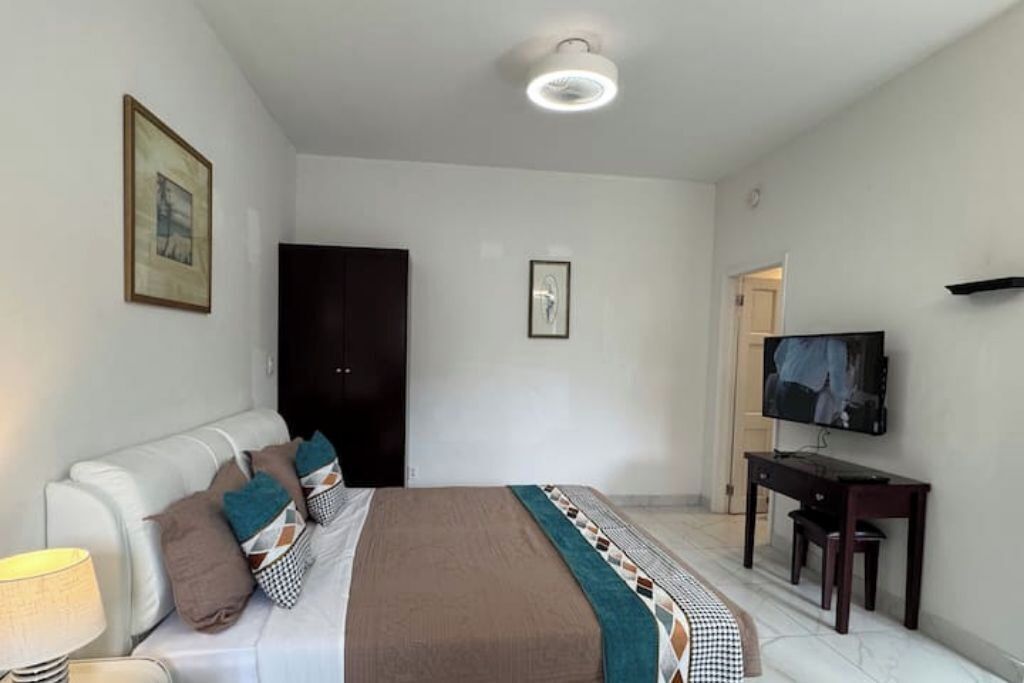 A modern hotel room with a bed, a desk with a computer, a television, and a door leading to another room.