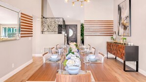 A dining room with a long wooden table set for six, surrounded by chairs. A chandelier hangs from the ceiling, and there is a decorative wall panel on the left.