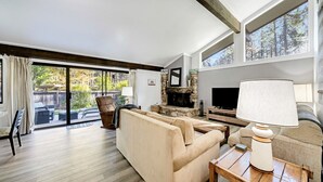 A spacious living room with a stone fireplace, a television, and a large window overlooking a backyard. - This light-filled alpine retreat blends rustic charm with modern style. (Graeagle)