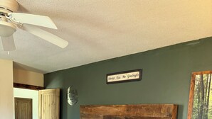 Un dormitorio con cabecera de madera, cama con ropa blanca, una mesita de noche de madera, un ventilador de techo y una puerta que da a otra habitación.