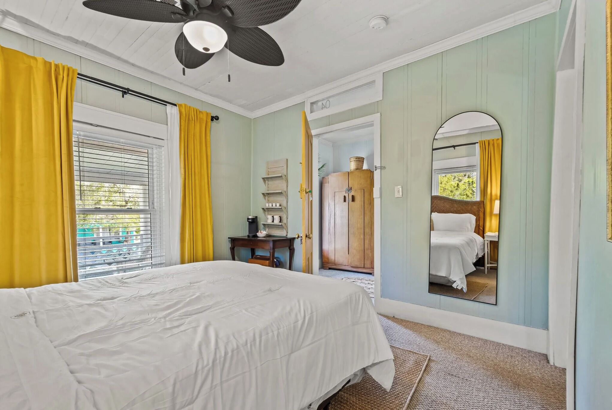 Une chambre à coucher avec un lit, un ventilateur de plafond, une fenêtre avec des rideaux jaunes, un miroir et une porte donnant sur une autre pièce.
