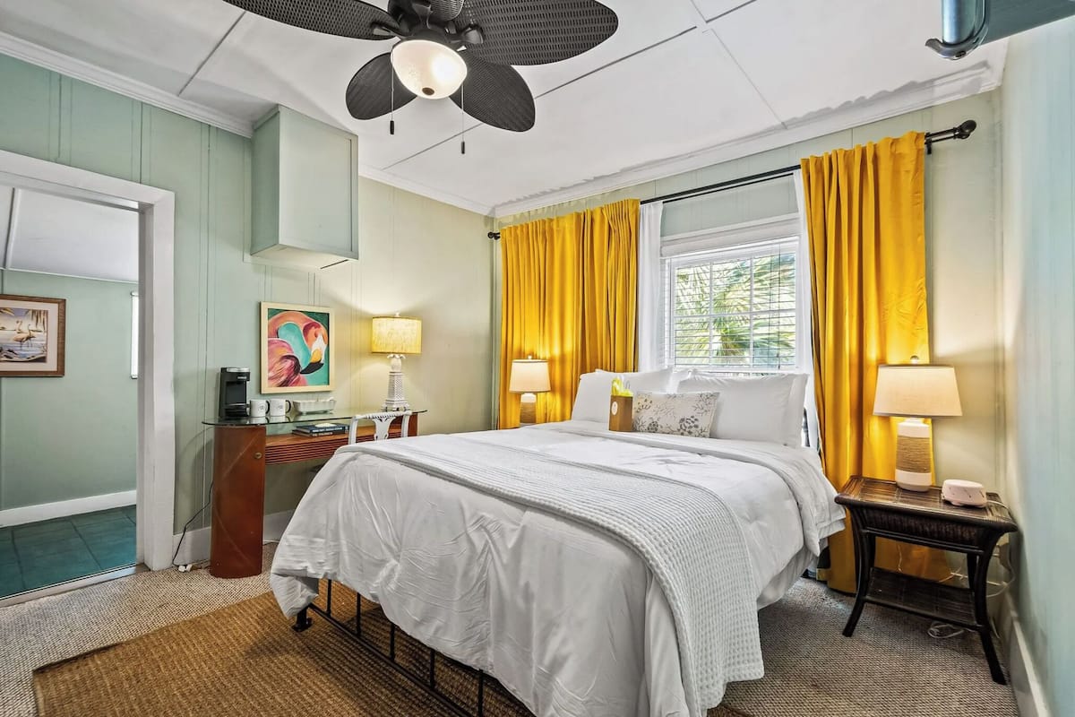 A hotel room with a bed, two bedside tables, a desk with a chair, a ceiling fan, and a window with yellow curtains.