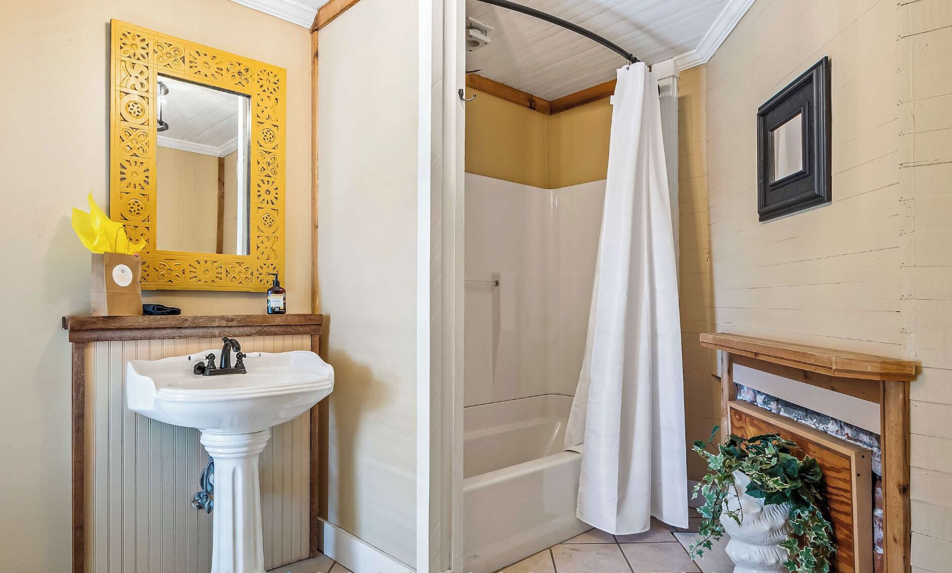 Un baño con un lavamanos blanco de pedestal, tocador de madera, espejo dorado ornamentado, ducha con cortina blanca y una pequeña planta en el suelo.