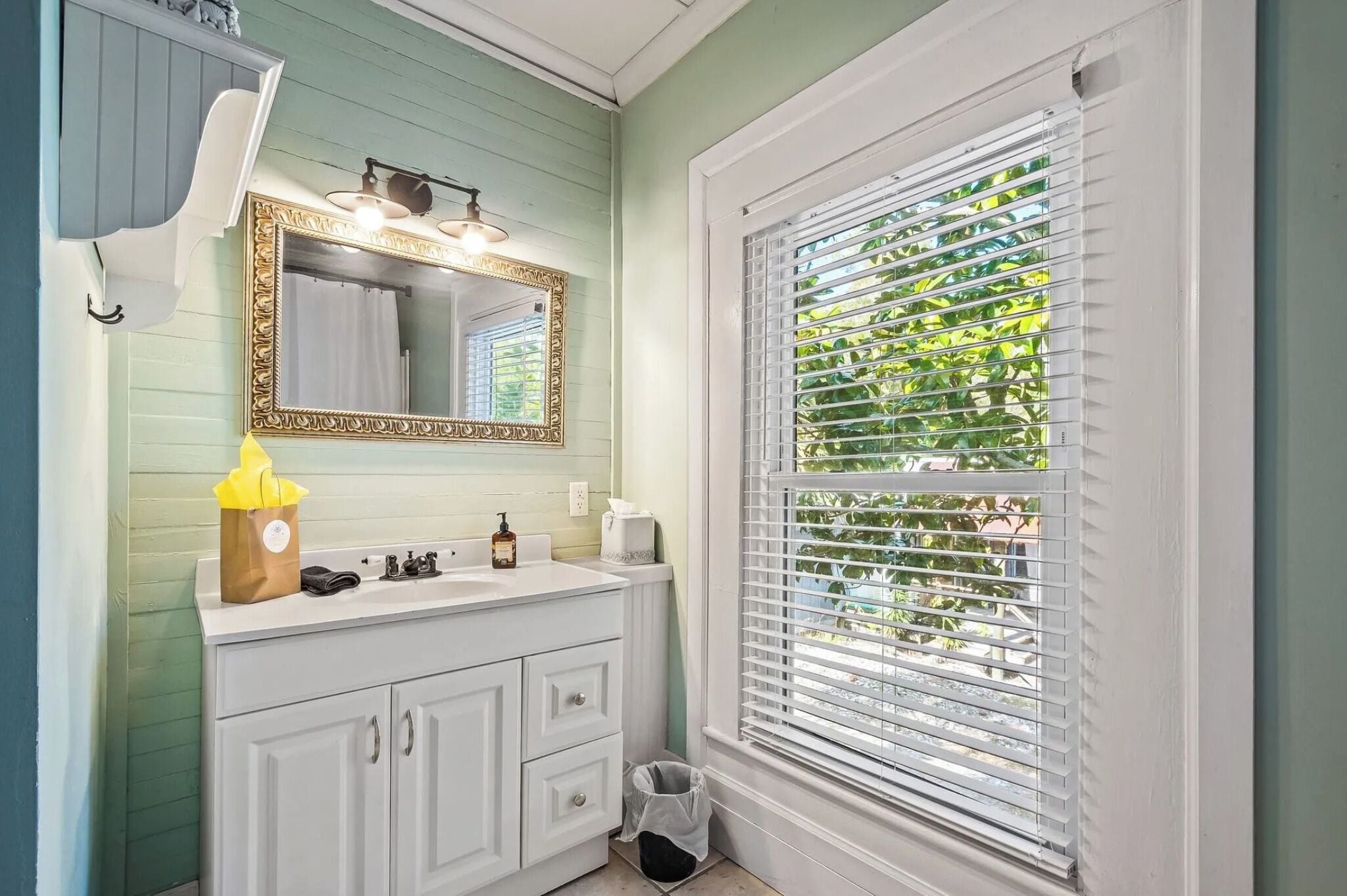 A bathroom with a white vanity, a gold-framed mirror, and a window with blinds.