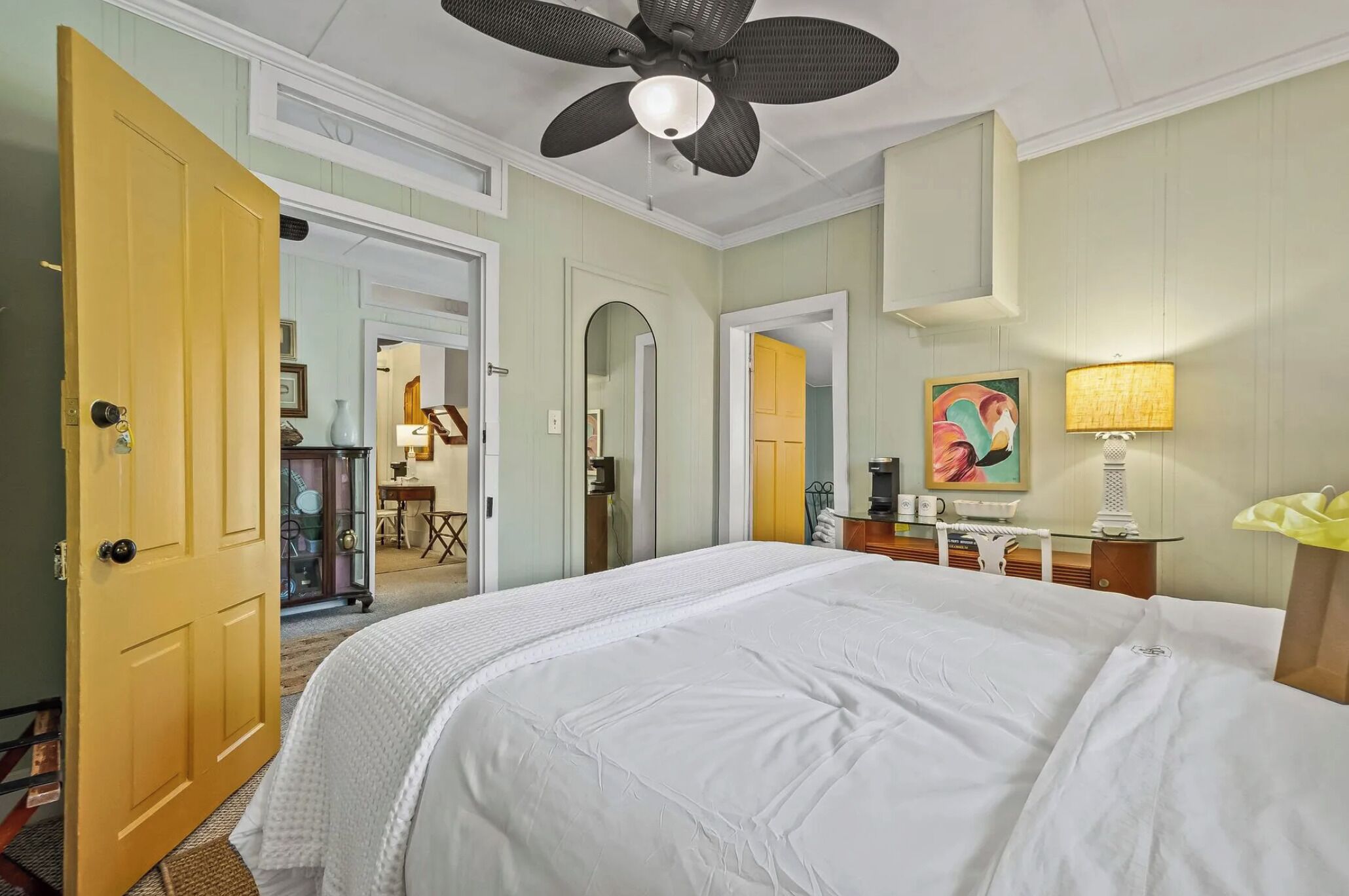 A hotel room with a large bed, a ceiling fan, a desk with a lamp, and a painting on the wall.