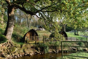 A rustic cabin with a wooden deck, surrounded by a natural landscape with a stream and trees. - Romantic Cosy Cabin with Hot Tub in Cumbria (Brampton)