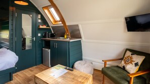 A compact room with a kitchenette, a bed, a wooden table with papers, a green armchair, and a television on the wall. - Romantic Cosy Cabin with Hot Tub in Cumbria (Brampton)