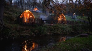 A row of wooden cabins with illuminated windows, a small stream, and a forested backdrop. - Romantic Cosy Cabin with Hot Tub in Cumbria (Brampton)