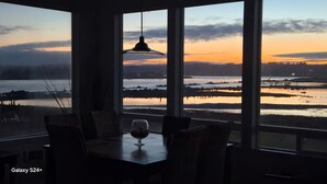 A dining area with a view of a sunset over a body of water, featuring a table with chairs and a wine glass. - The Coquille River Retreat (Bandon)