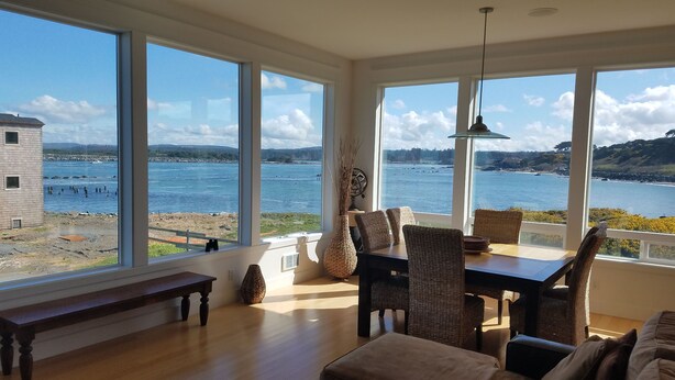A room with large windows overlooking a body of water, a dining area with wicker furniture, and a wooden bench. - The Coquille River Retreat (Bandon)