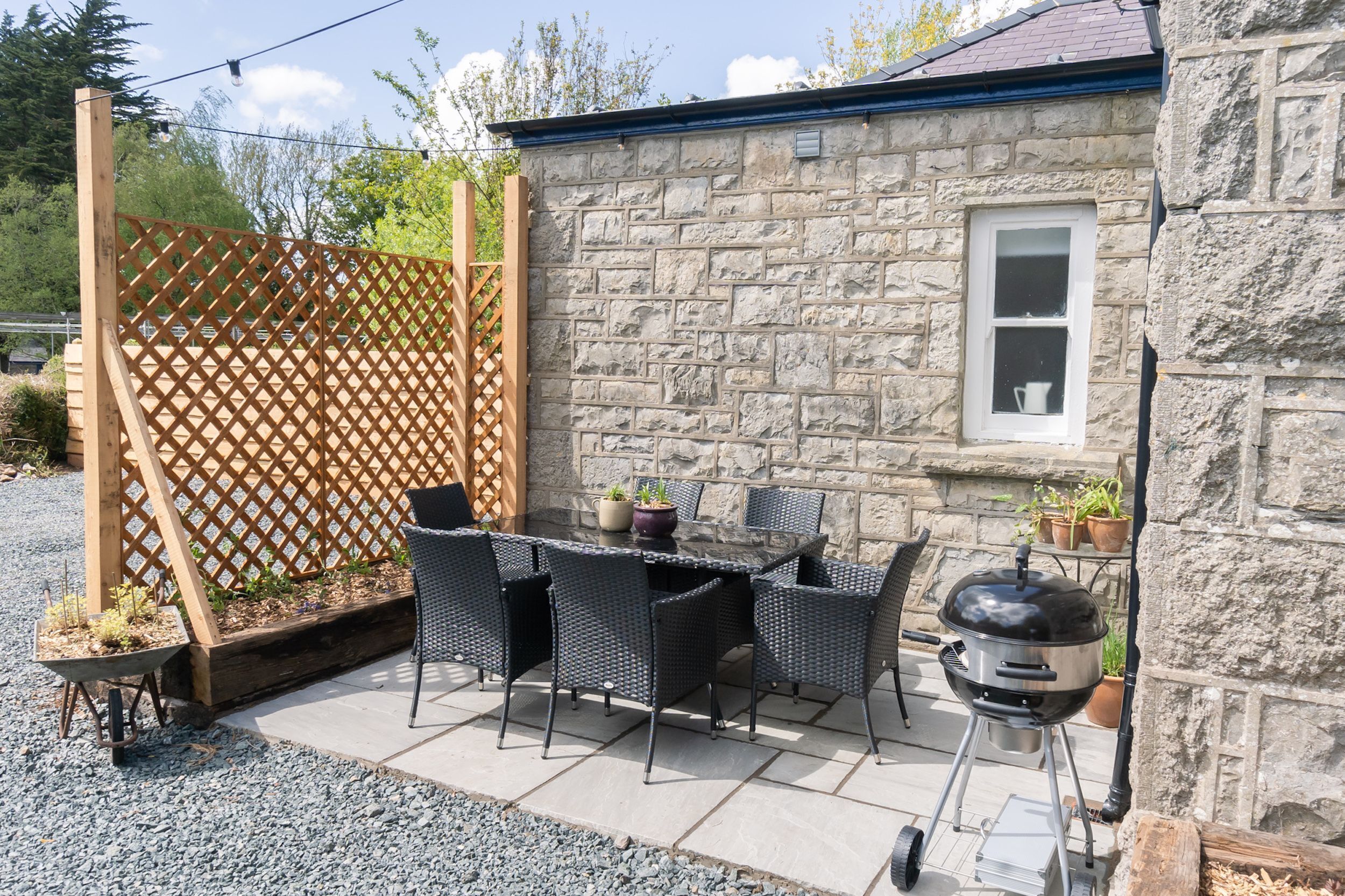 En udendørs terrasse med stenvæg, et træhegn, et bord med stole, grill og plantekasser.