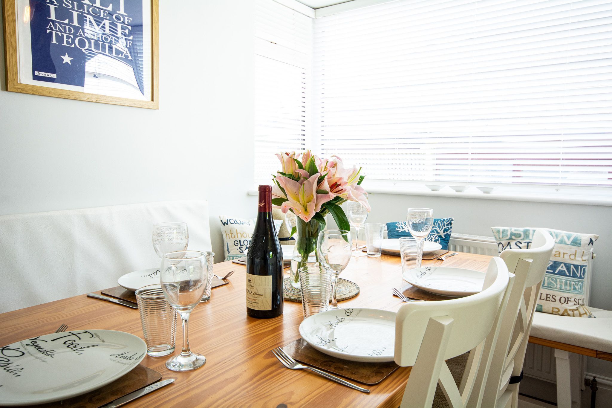 Une table à manger dressée avec des assiettes, des verres et une bouteille de vin, avec une fenêtre et des stores en arrière-plan.