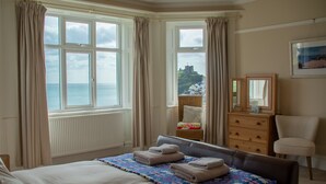 Una camera d'albergo con un letto, una cassettiera, una sedia e vista sul mare e su un castello attraverso ampie finestre.