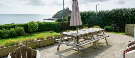 An outdoor patio with a picnic table, chairs, and an umbrella. A view of the sea and a hill with a castle-like structure is visible in the background.