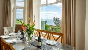 Tavolo apparecchiato per due, con bottiglia di vino, bicchieri e un vaso di fiori. Il tavolo è posizionato vicino a una finestra con vista sul mare e sul verde.