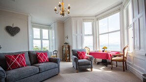 A living room with a grey sofa, a small armchair, and a dining table with a red tablecloth. - Llety - sleeps 2 guests  in 1 bedroom (Criccieth)