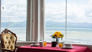 A dining table with a red tablecloth, two cups, a vase with sunflowers, and a view of the sea and mountains through large windows. - Llety - sleeps 2 guests  in 1 bedroom (Criccieth)