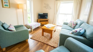 Un salon avec deux canapés, une table basse, un téléviseur posé sur un meuble en bois et une grande fenêtre avec des rideaux.