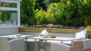An outdoor seating area with wicker furniture, a table, and a window overlooking a garden with rocks and plants. - Goleufryn - sleeps 20 guests  in 7 bedrooms (Abersoch)