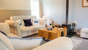 A cozy living room with a sofa, a coffee table with a book and wine, and a wood-burning stove. - Cosy Luxury Cottage - Hot Tub - Private Estate (Rhosneigr)