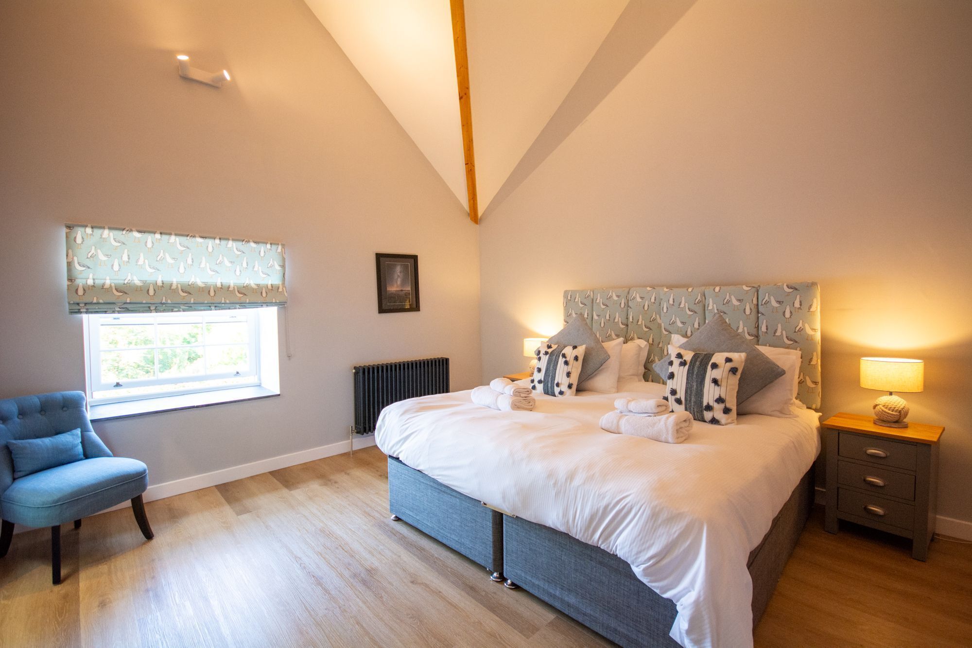 A bedroom with a bed, a chair, a bedside table, a lamp, a window with curtains, and a radiator.