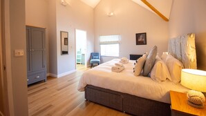 A bedroom with a large bed, a bedside table with a lamp, a wardrobe, and a window with a view. - Stylish Cottage - Hot Tub - Garden - Near Beach (Rhosneigr)