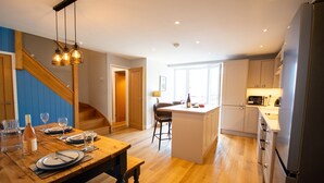 A modern kitchen with wooden flooring, a dining area with a wooden table set for four, and a staircase leading to another level. - Stylish Cottage - Hot Tub - Garden - Near Beach (Rhosneigr)